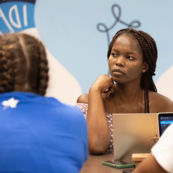 A Black female student listens intently and looks to the right while seated at a table with another student in the foreground with a blue shirt with back to the camera. 